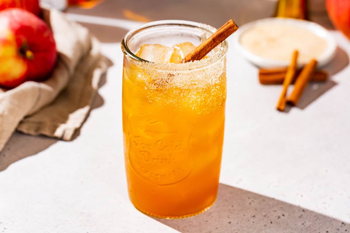 Large tumbler style cocktail glass with a Fireball Apple Cider cocktail over ice. The drink is garnished with a cinnamon stick and cinnamon sugar on the rim.