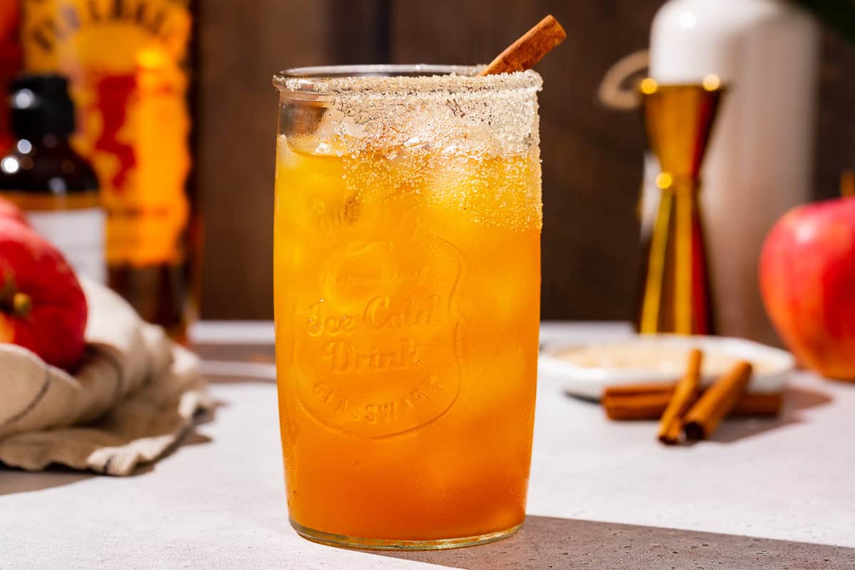 Side view of a Fireball Apple Cider cocktail. Fireball whiskey is in the background along with a jigger, apples and cinnamon sticks.