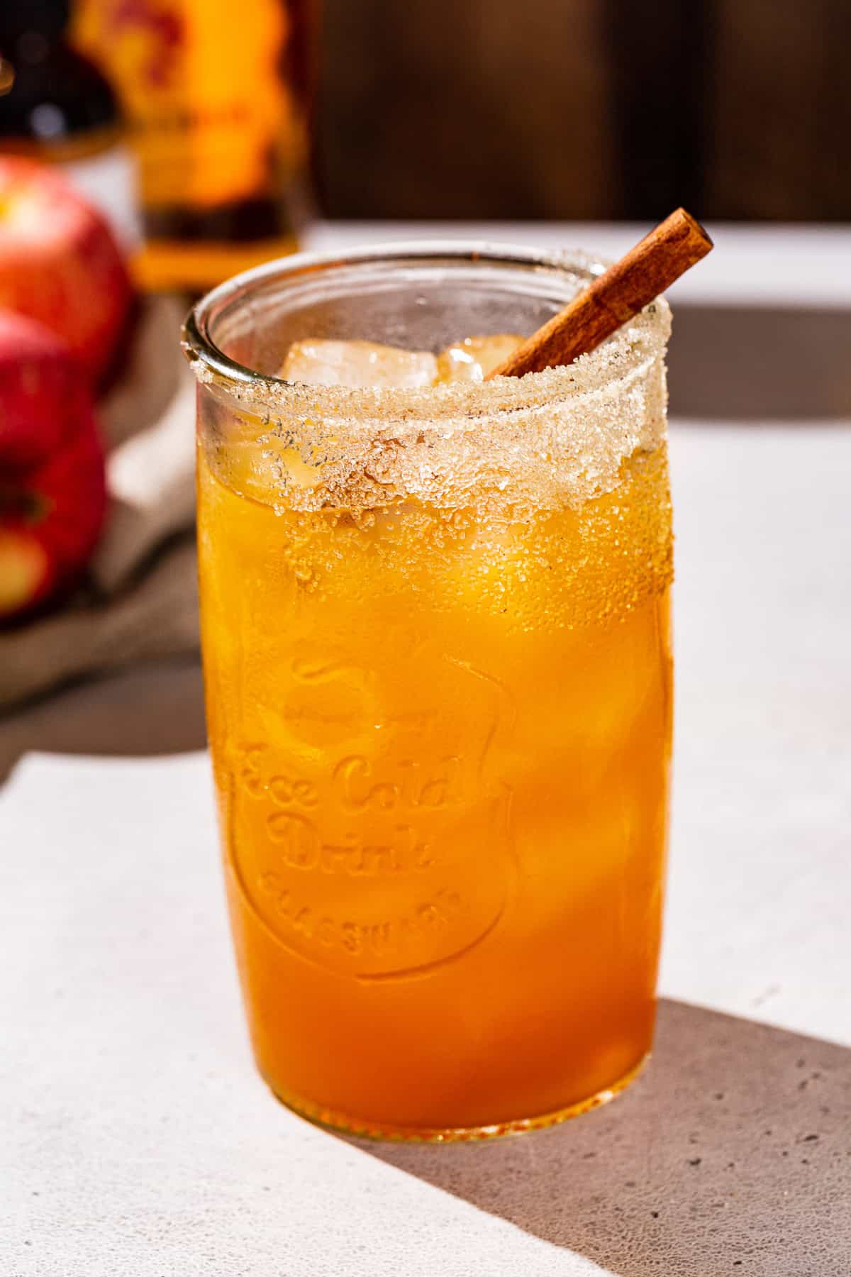 Fireball whiskey and apple cider cocktail with a cinnamon stick garnish. Apples and fireball bottle are slightly visible in the background.