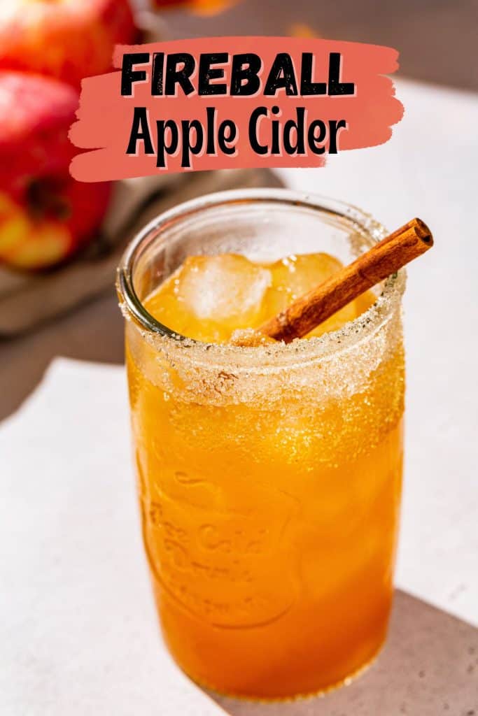 Slightly overhead photo of a Fireball Apple Cider cocktail over ice. The serving glass has cinnamon sugar on the rim. Text above the drink says “Fireball Apple Cider” in bold letters.