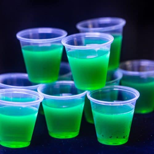 Glow in the dark jello shots shown glowing under a blacklight with a black background. The shots are green in color.