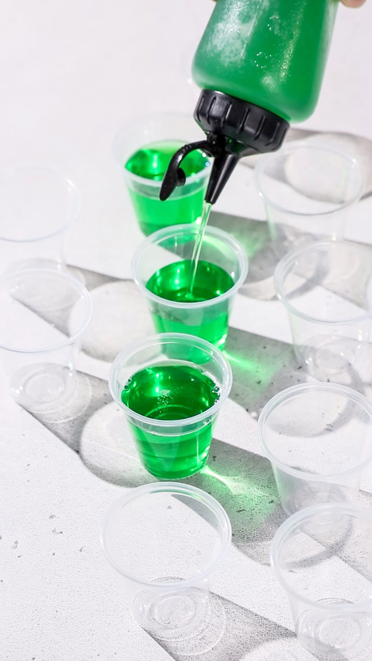 Hand using a squeeze bottle filled with the jello shot mixture to fill up plastic cups before chilling them in the fridge.