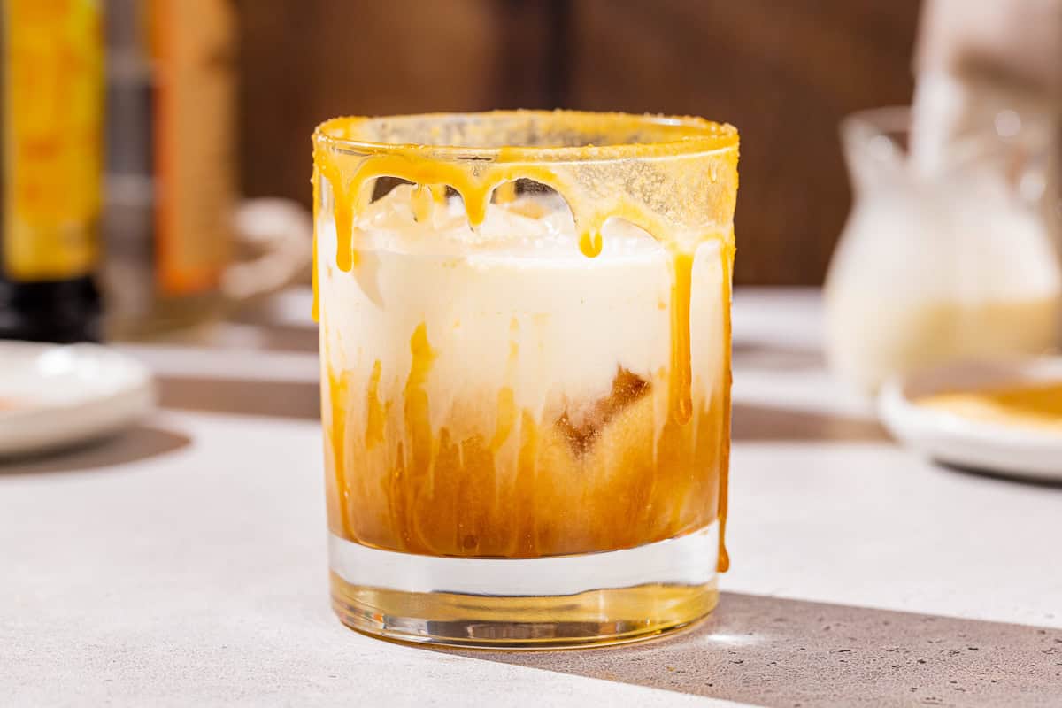 Maple Caramel white Russian cocktail in an old fashioned glass. The cocktail has a brown layer on the bottom and white cream layer on top. In the background are ingredients used to make the drink.