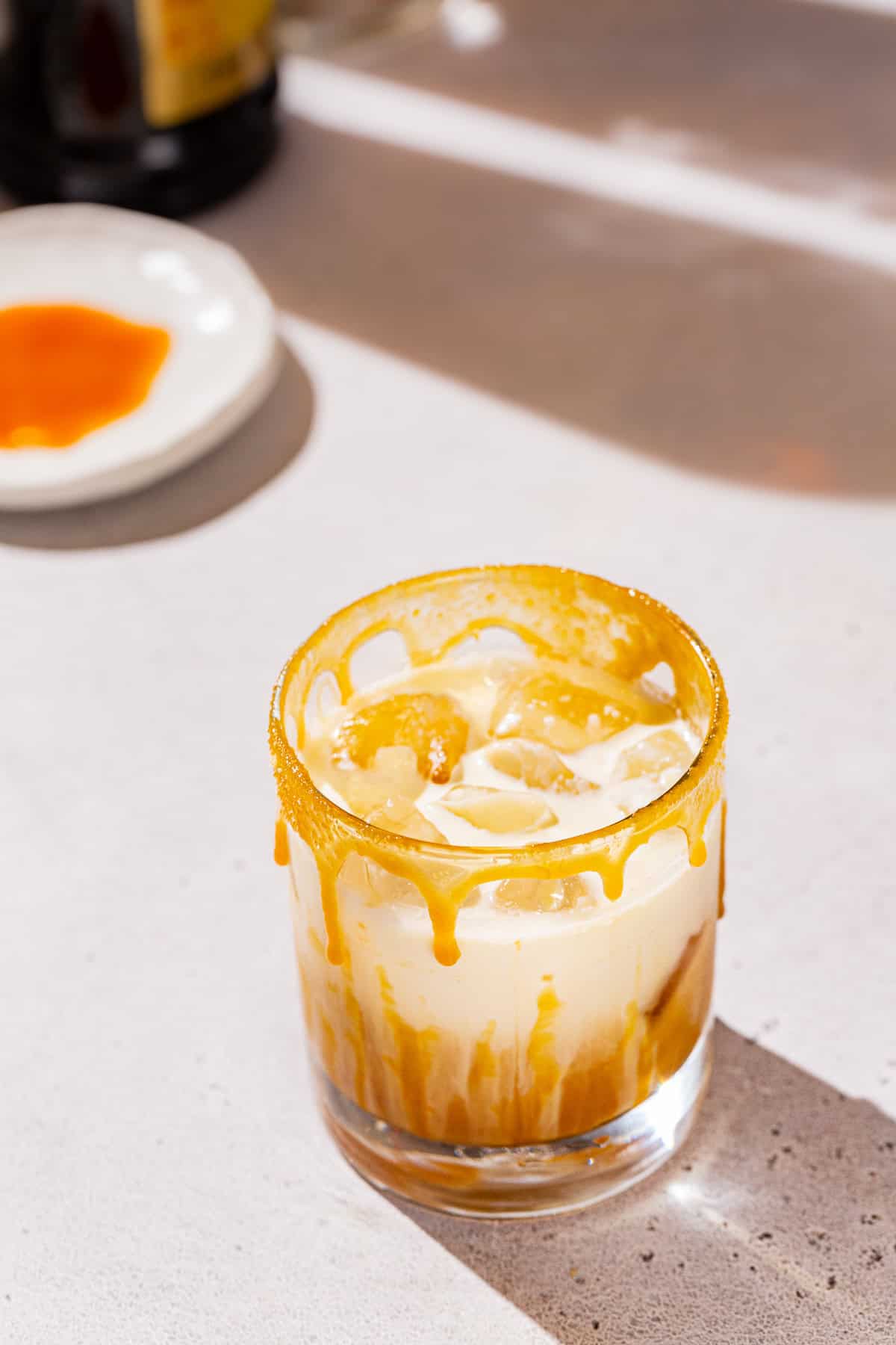 Caramel Maple white Russian cocktail with a caramel drizzle on the side of the glass. In the background is a dish of caramel sauce and a bottle of Kahlua.