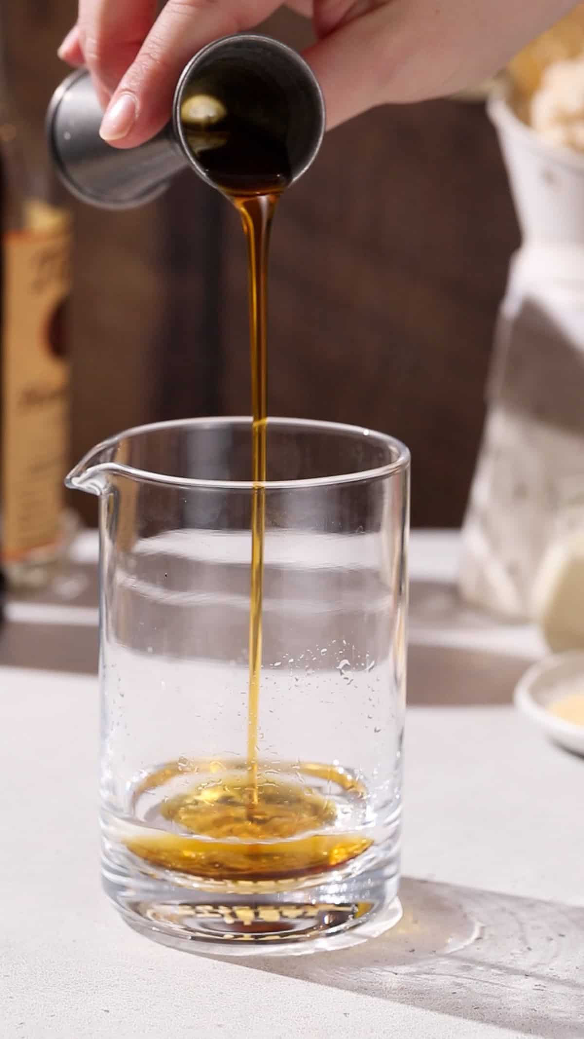 Hand pouring maple syrup into a cocktail mixing glass.