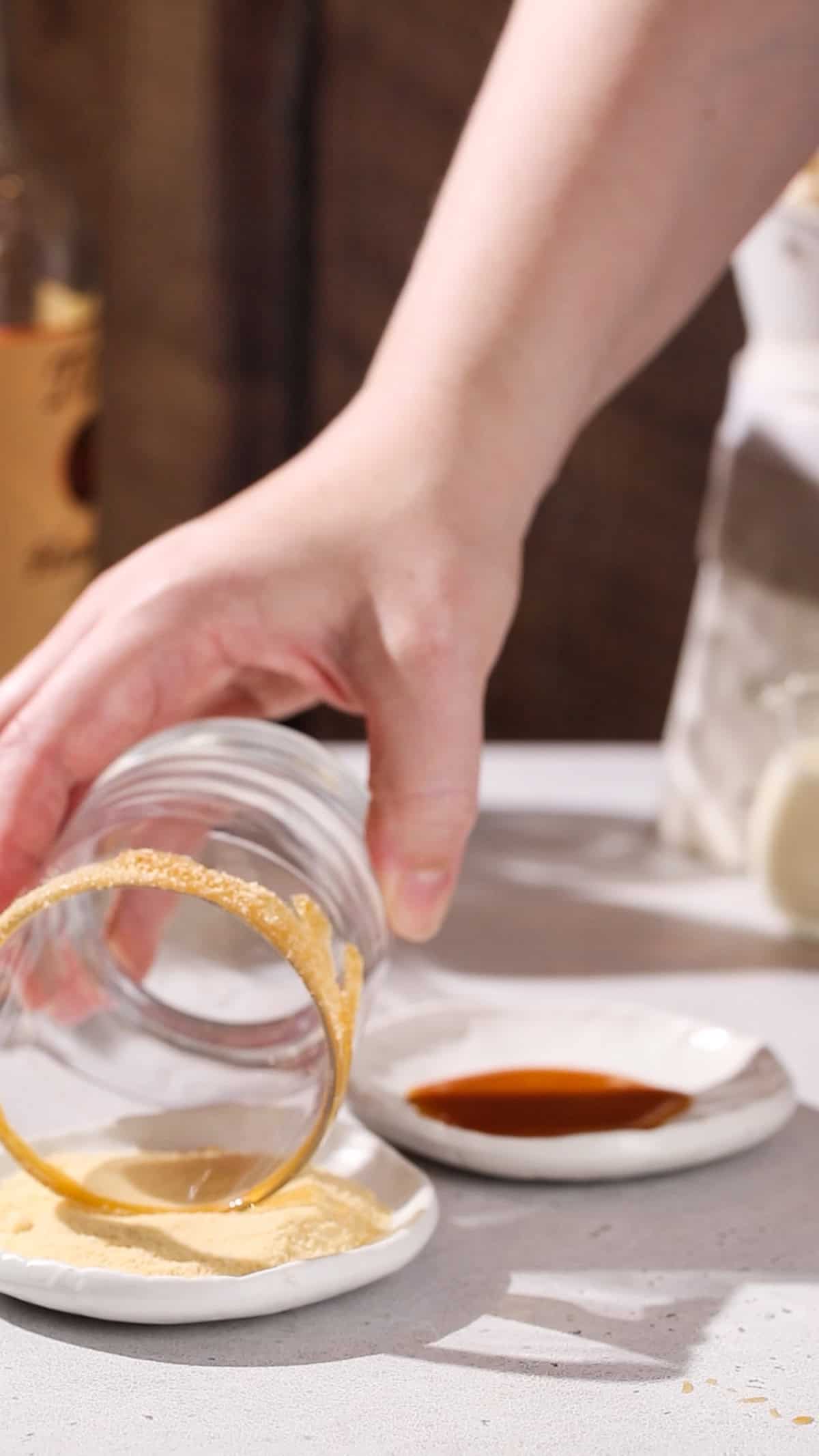 Hand dipping a caramel-coated cocktail glass rim into a dish of maple sugar.