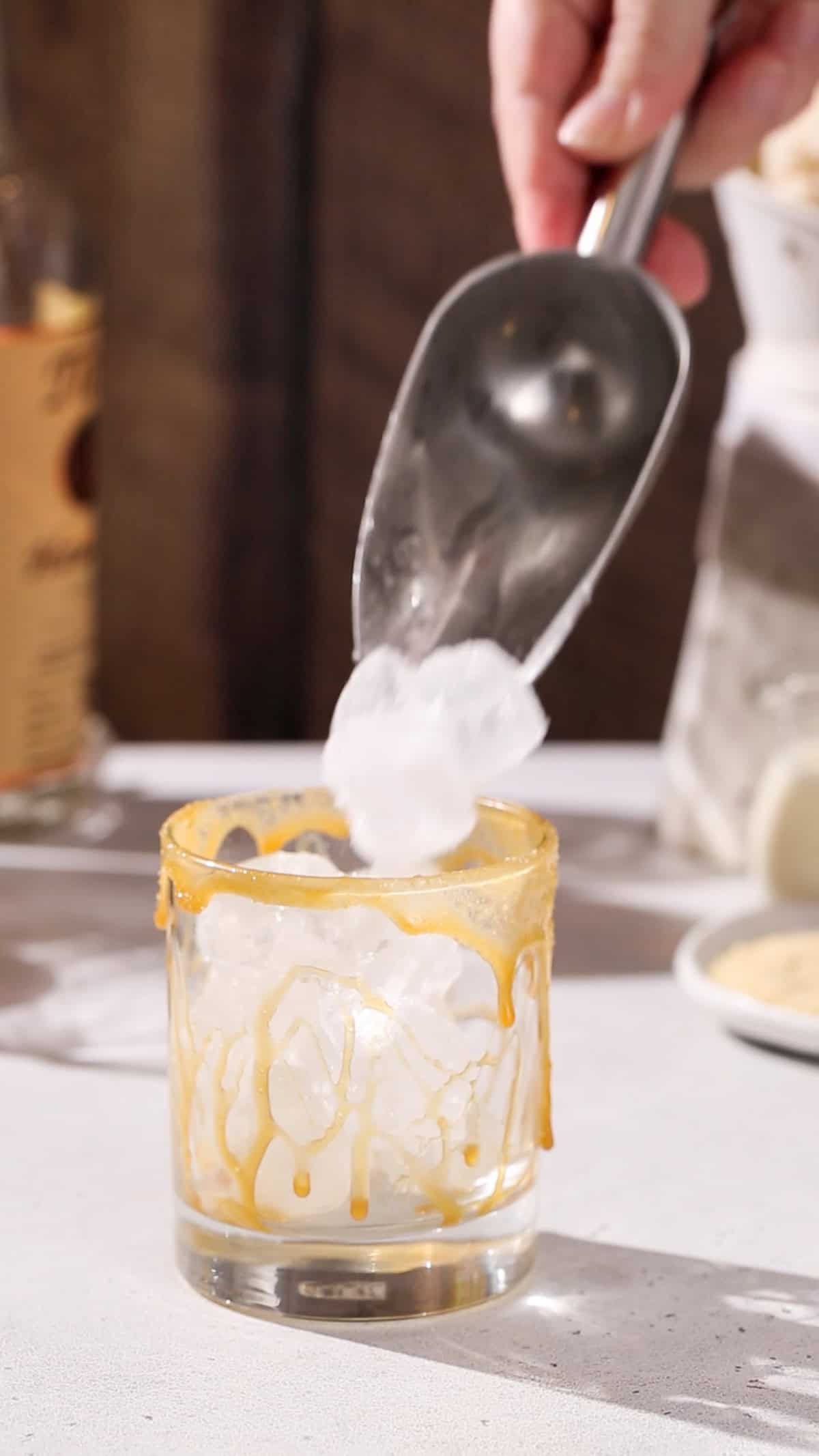 Hand adding ice to a caramel-drizzled cocktail glass.