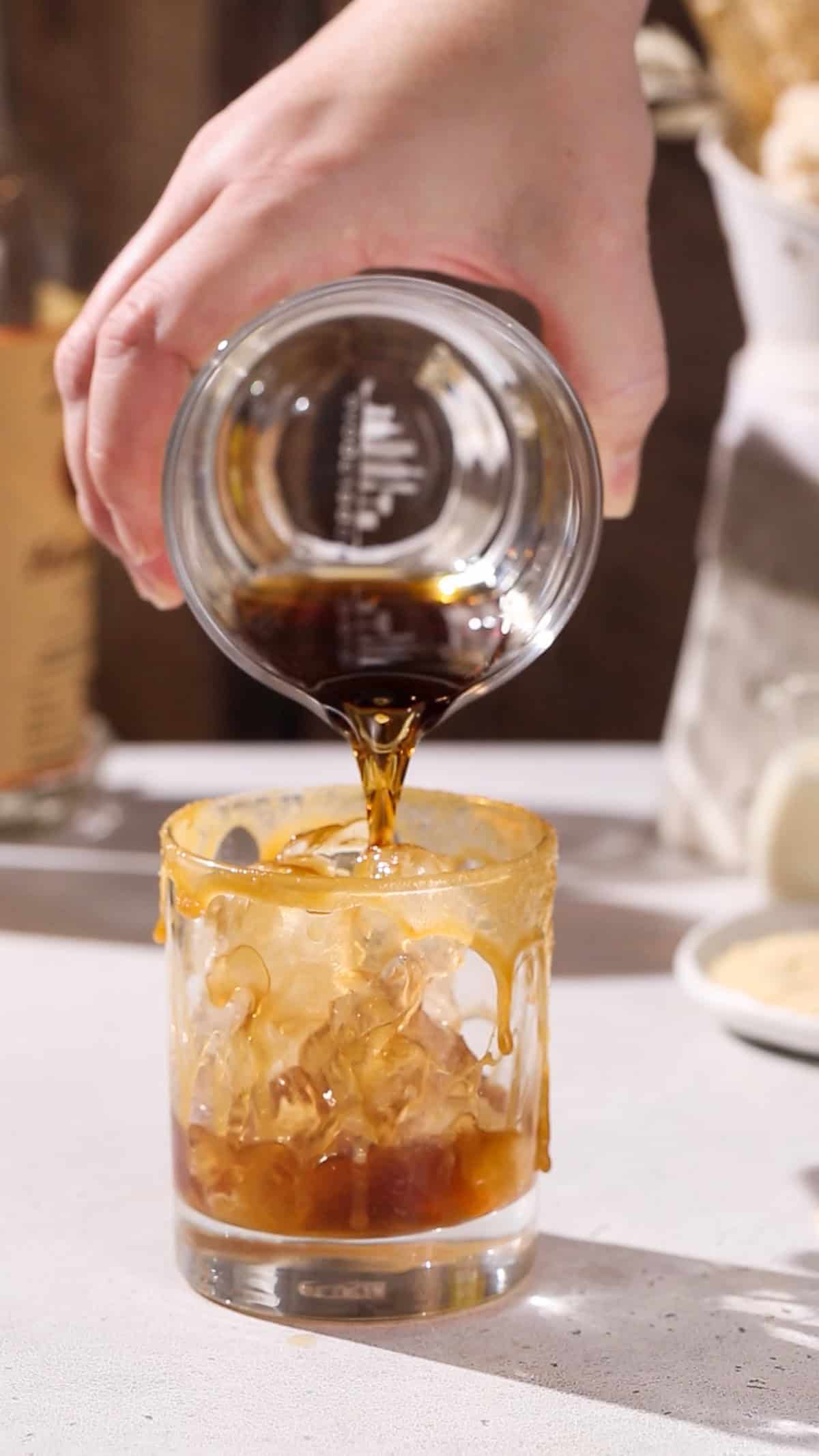 Hand pouring brown liquid into a cocktail glass filled with ice and caramel sauce.