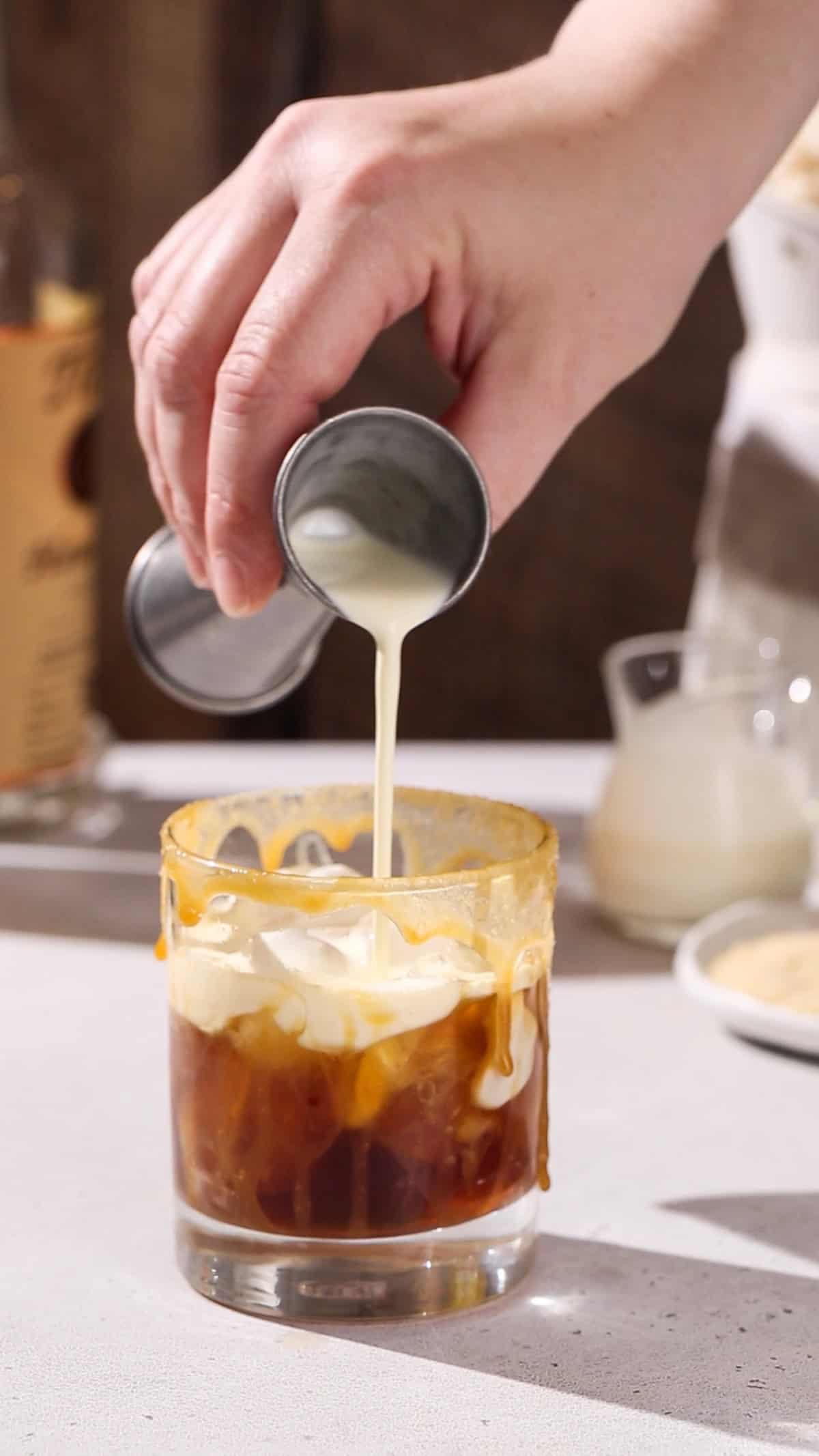 Hand pouring a layer of white cream on top of a brown cocktail in a glass with a caramel drip on the side.