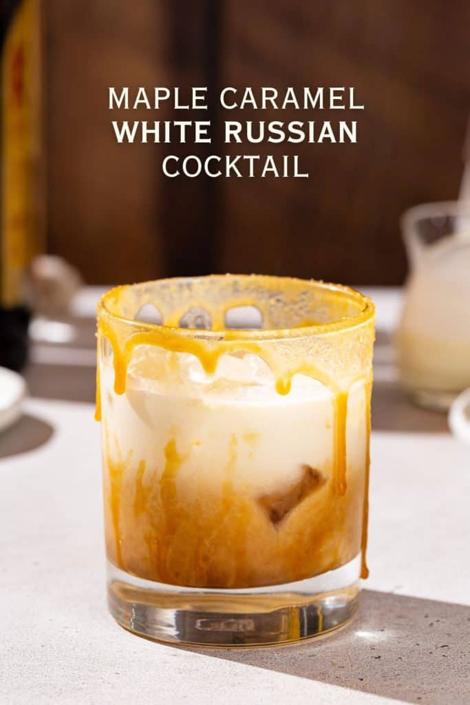 Side view of a Maple Caramel White Russian drink on a countertop with ingredients in the background of the image. Text above the drink says “Maple Caramel White Russian cocktail”.