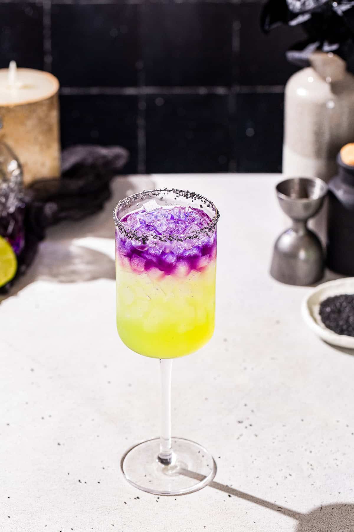 Witches’ Brew cocktail on a countertop with ingredients and spooky looking props around it. The drink is green on the bottom and purple on the top.