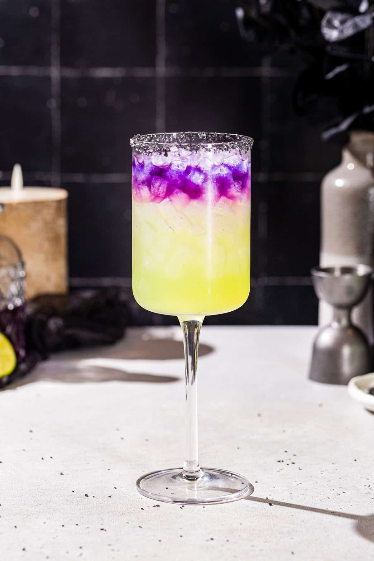 Side view of a Witches Brew cocktail with a green bottom and purple layer on top in a tall wine glass. There are bar tools and a candle in the background.