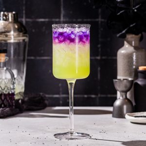 Side view of a Witches’ Brew cocktail with a green layer on the bottom and purple on the top. The drink is in a tall wine glass with a black salt rim. Bar tools are seen in the background.