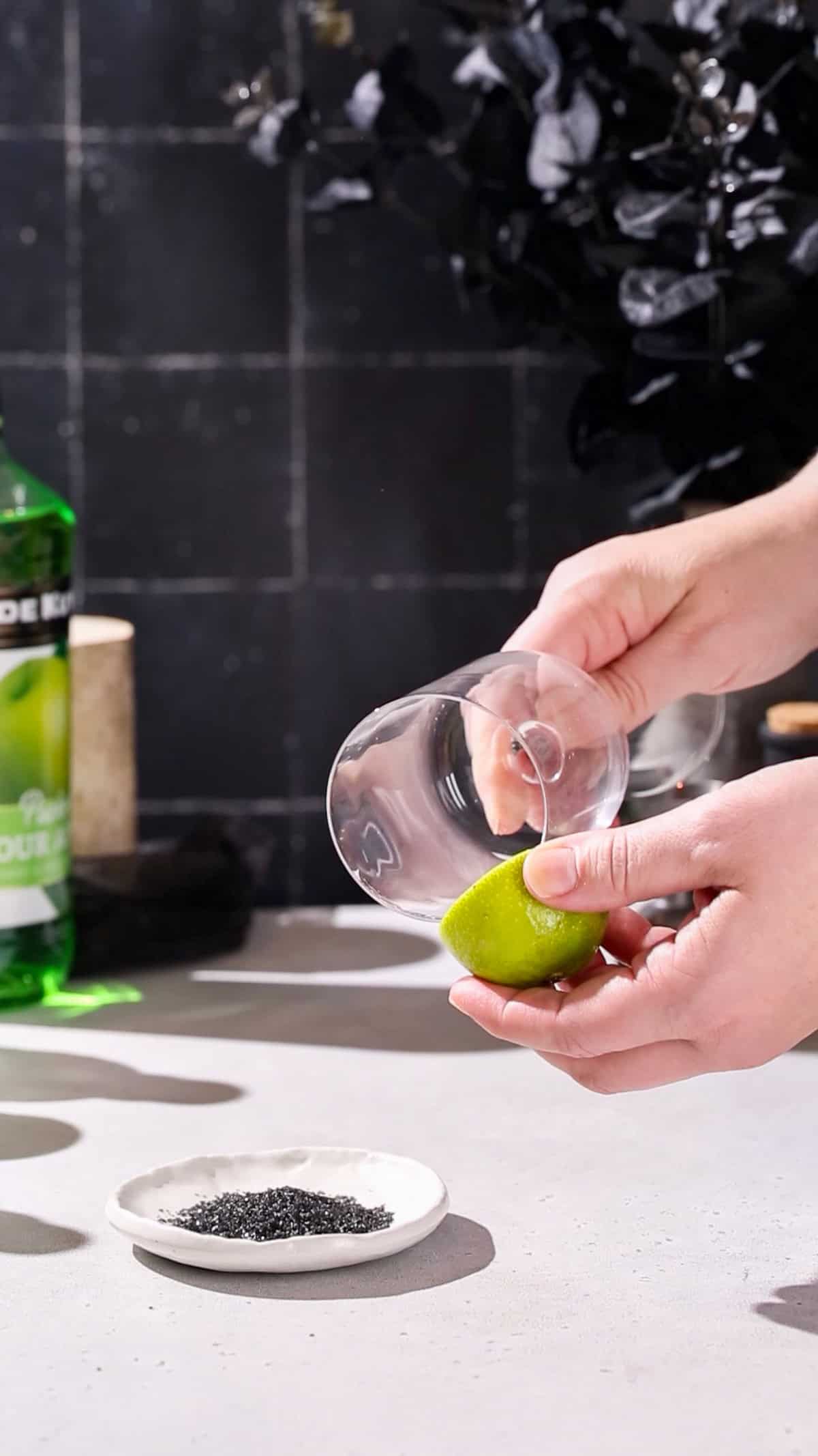 Hand using a lime to wet the rim of a tall wine glass with straight sides.