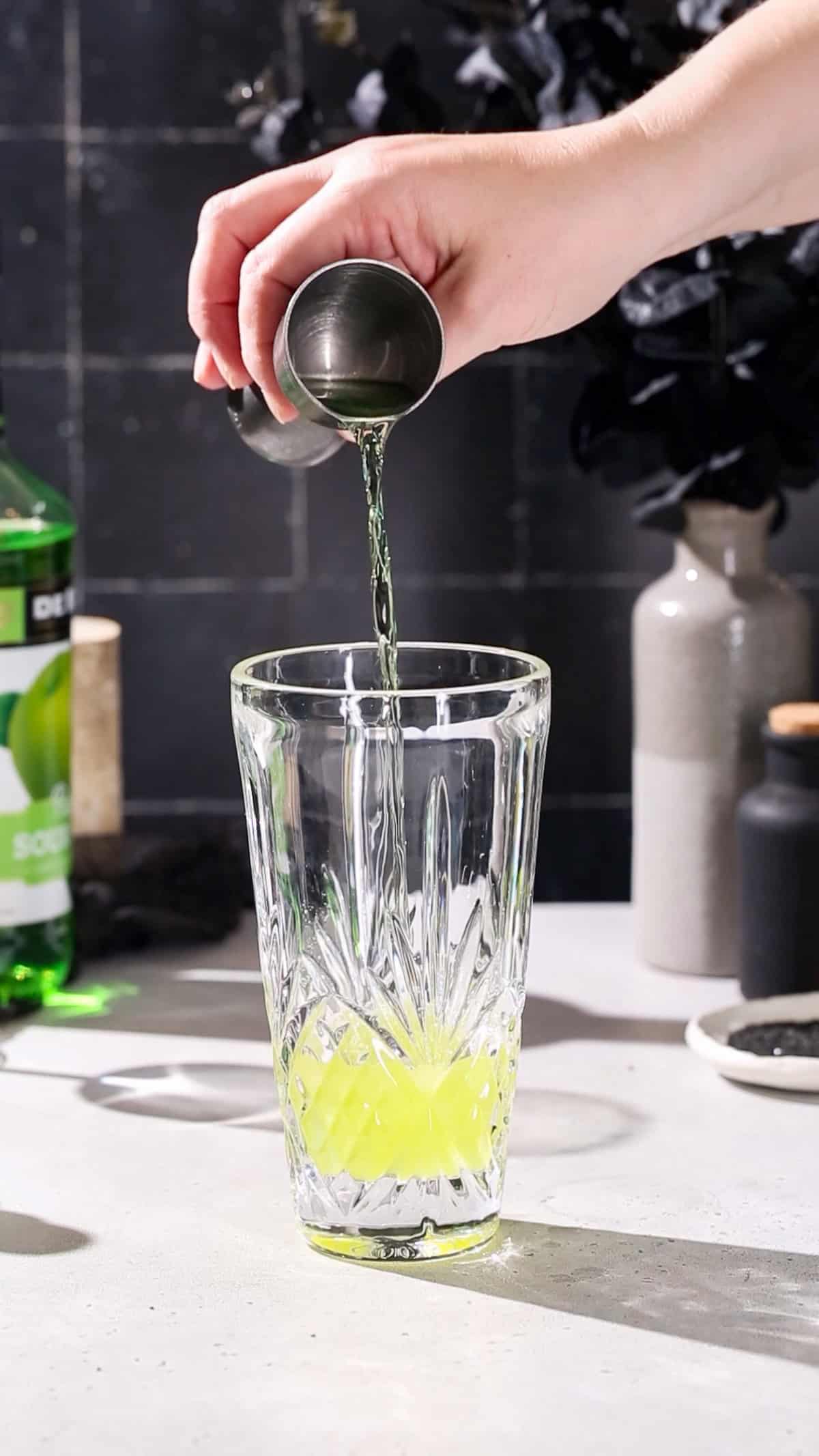 Hand adding sour apple pucker liqueur to a cocktail shaker.