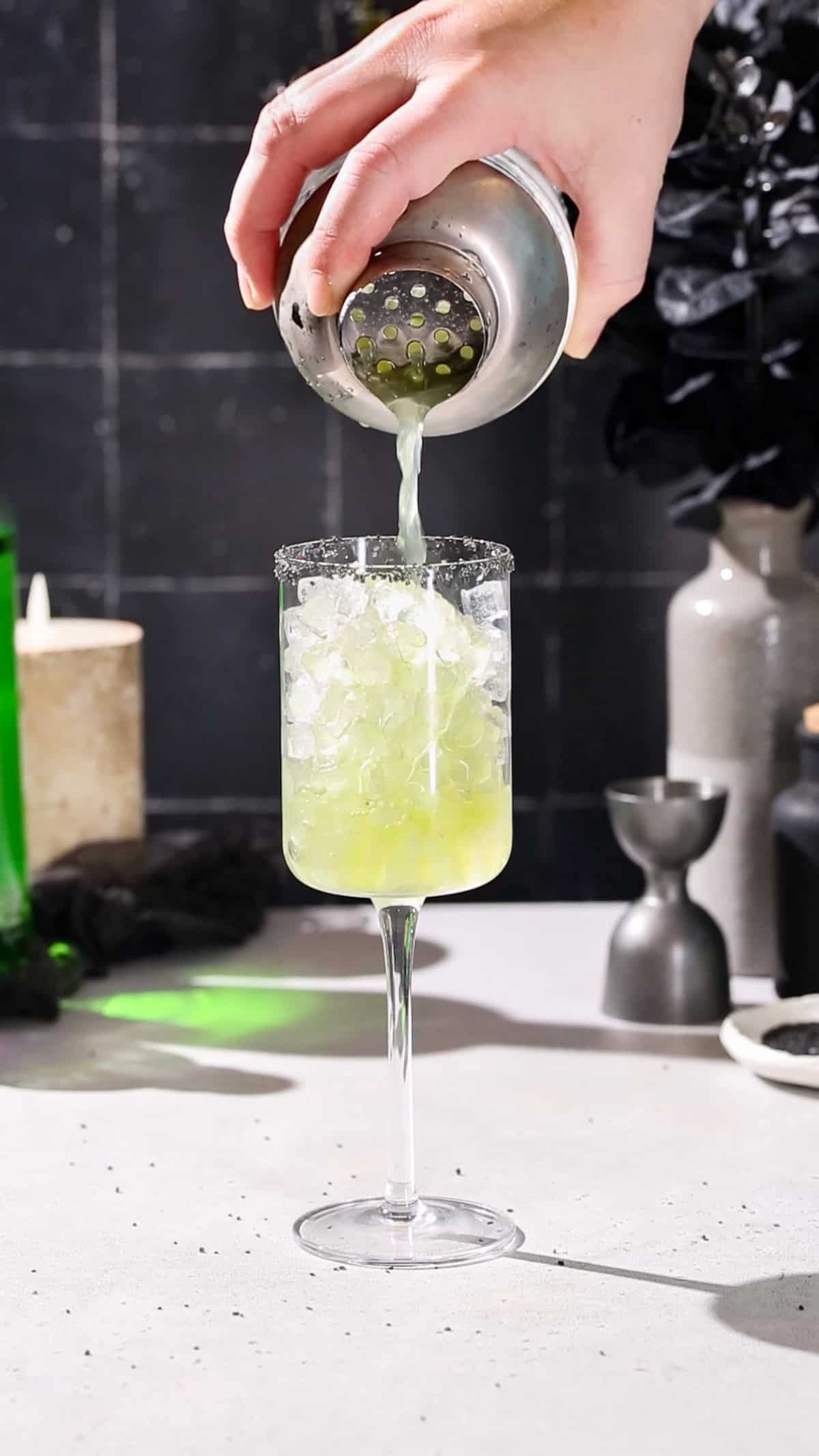 Hand straining a green cocktail in to a wine glass filled with pebble ice.