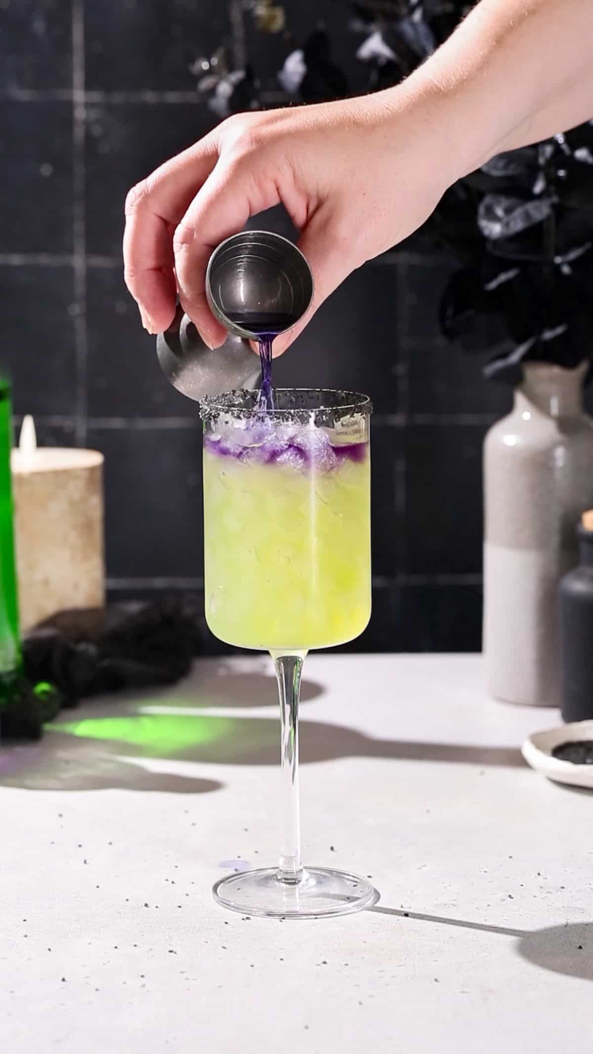 Hand pouring purple butterfly pea tequila onto the top of a green cocktail.