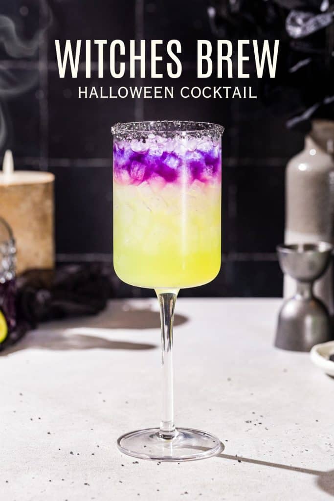 Purple and green Witches brew cocktail in a tall wine glass with a black salt rim. Text above the drink says “Witches’ Brew halloween cocktail”.