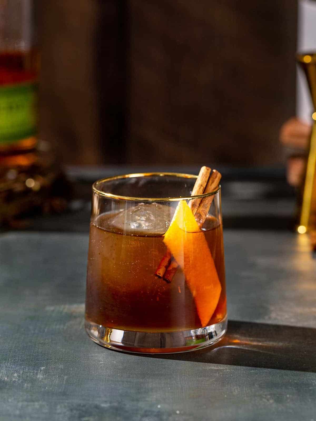 Side view of a Gingerbread Old Fashioned cocktail in an old fashioned style glass, also called a lowball glass. The drink is brown and has a large sphere of ice in the glass along with an orange peel and a cinnamon stick garnish. In the background are a bottle of Bulleit rye whiskey and a gold jigger.