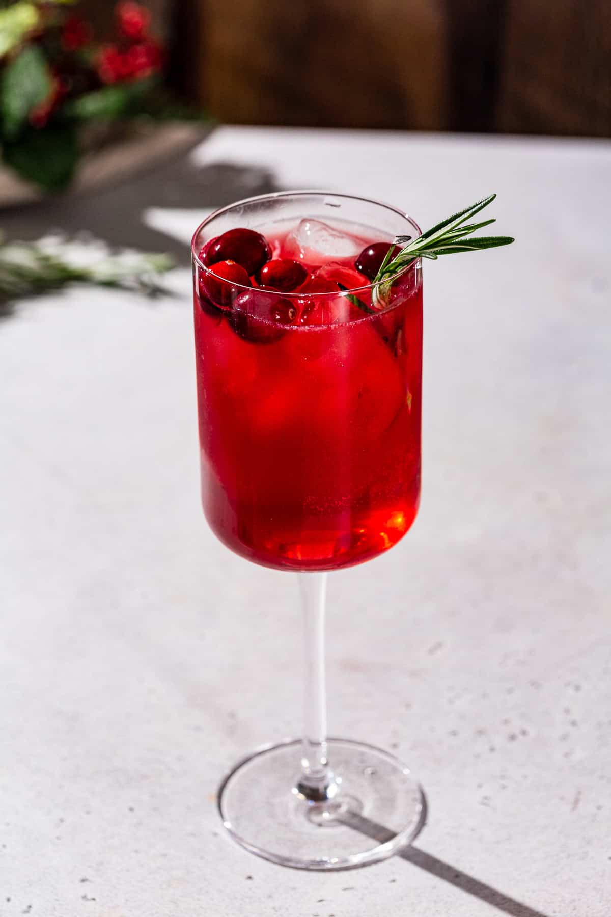 Overhead view of a Holiday Gin & Tonic cocktail. The drink is red and garnished with fresh cranberries and rosemary. Christmas decor is in the background.