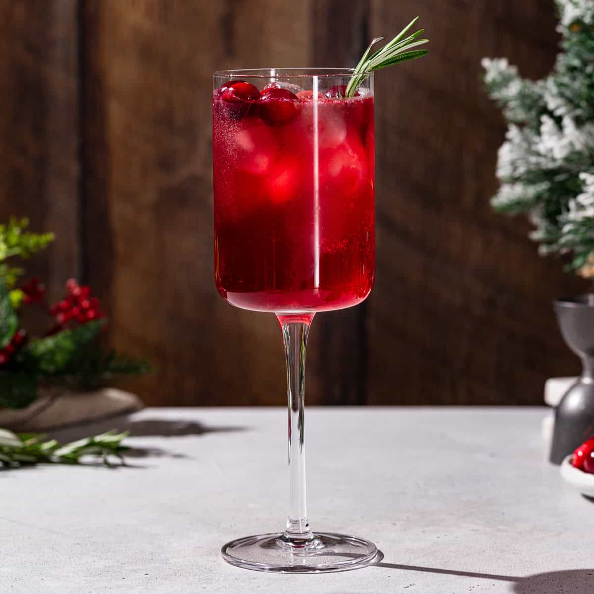 Side view of a Holiday gin and tonic cocktail in a square wine glass. The drink is red and garnished with cranberries and fresh rosemary. In the background is some Christmas evergreen decorations.