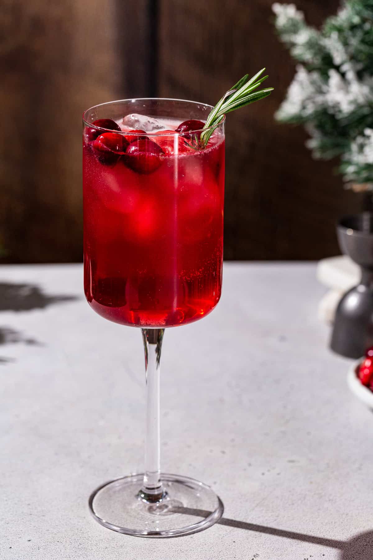 Side view of a Holiday gin and tonic cocktail in a square wine glass. The drink is red and garnished with cranberries and fresh rosemary.