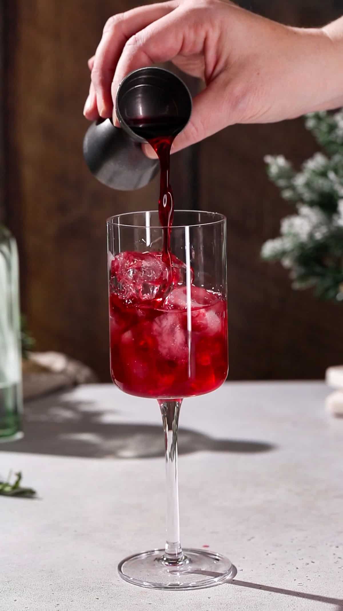 Hand adding cranberry juice to a cocktail glass filled with ice.