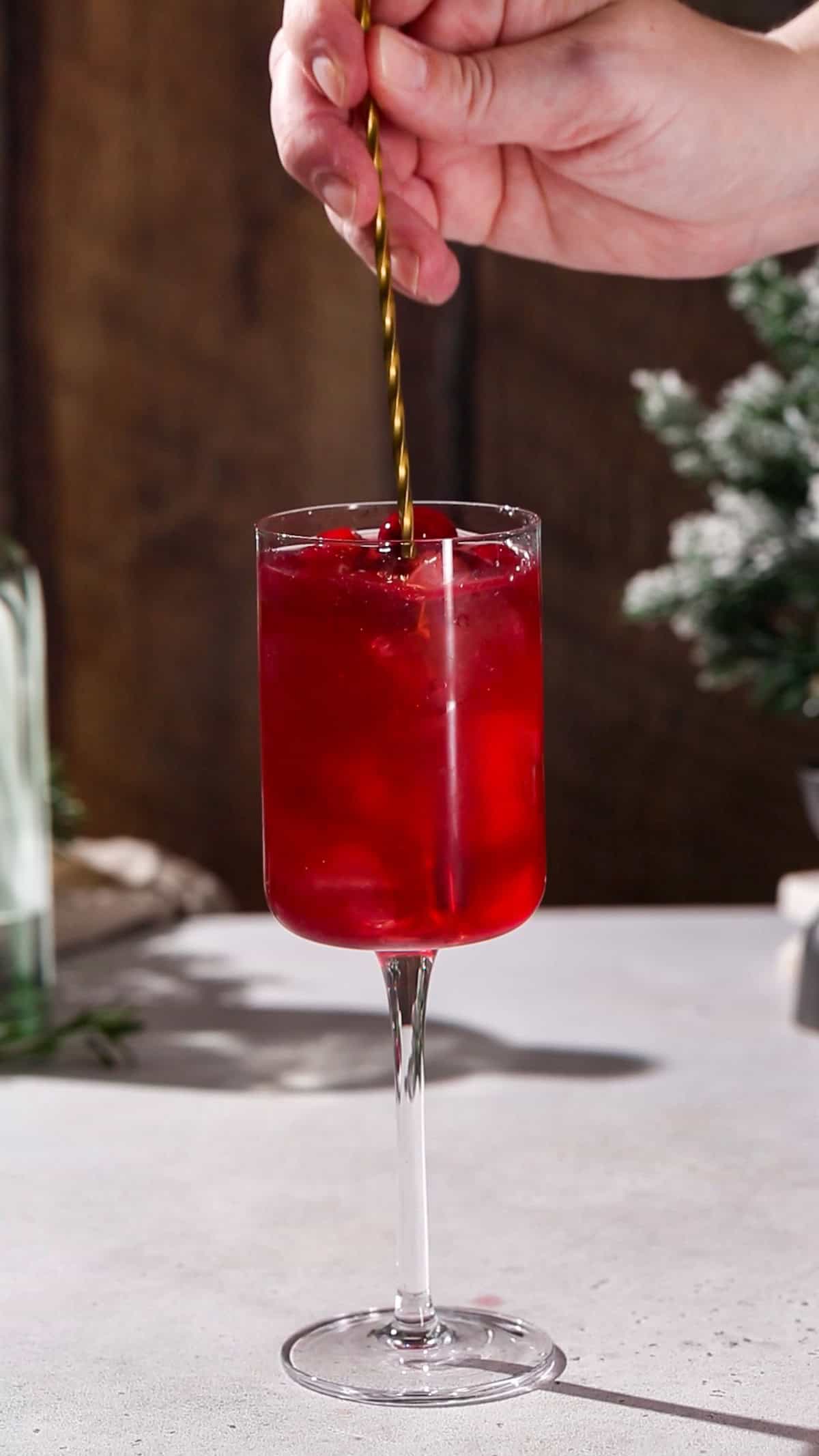 Hand using a long bar spoon to stir a red cocktail.