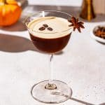 Pumpkin Spice Espresso Martini cocktail on a countertop garnished with three coffee beans and a star anise. Pumpkins and bar tools are in the background.
