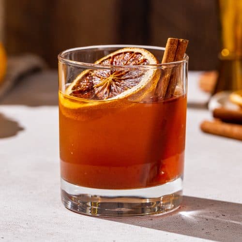 Close up of a Pumpkin Spice Old Fashioned cocktail in an old fashioned glass, garnished with a dried orange slice and cinnamon stick.