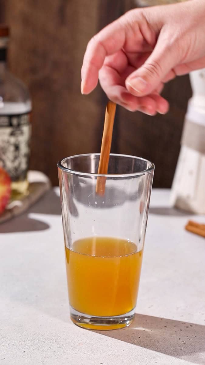 Hand adding a cinnamon stick to a cocktail shaker.