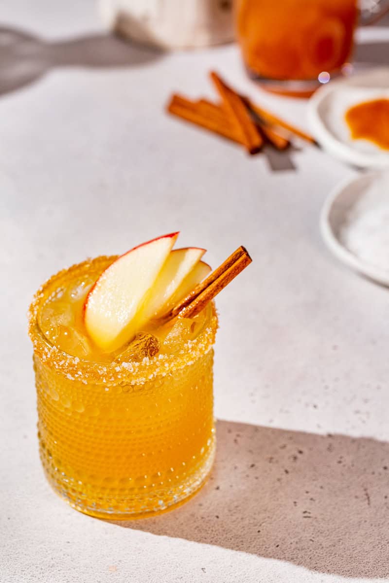 Overhead view of a Thanksgiving Margarita with apple slices and cinnamon stick garnish. There are cinnamon sticks and the dishes used to add the caramel rim to the glass in the background.