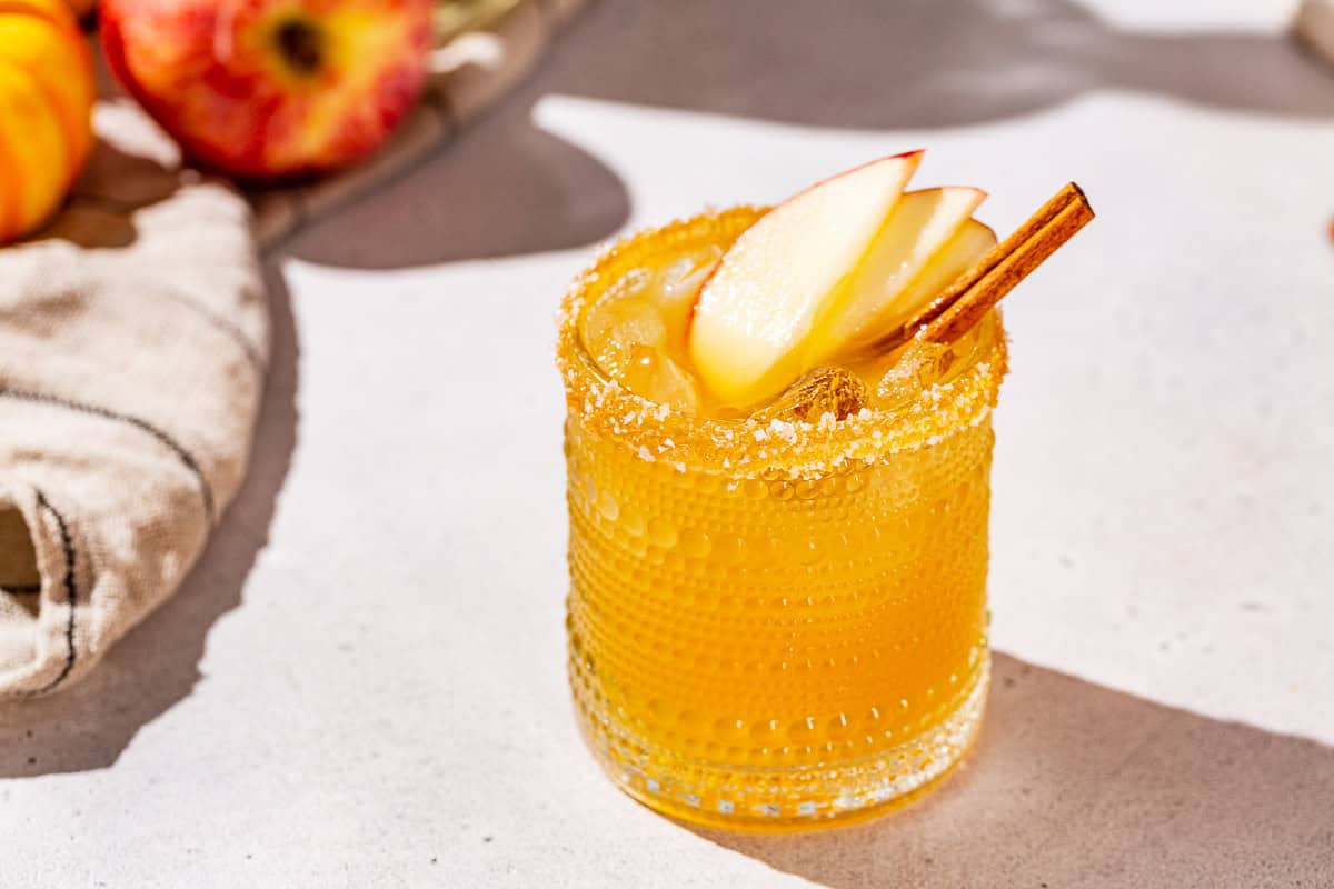 Apple cider Thanksgiving Margarita with apple slice and cinnamon stick garnish on a countertop. A whole apple is seen in the background.