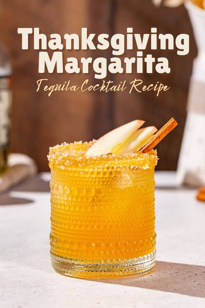 Side view of a Thanksgiving Margarita in a hobnail glass with apple slices and cinnamon stick garnish. Text above the drink says "Thanksgiving Margarita tequila cocktail recipe".