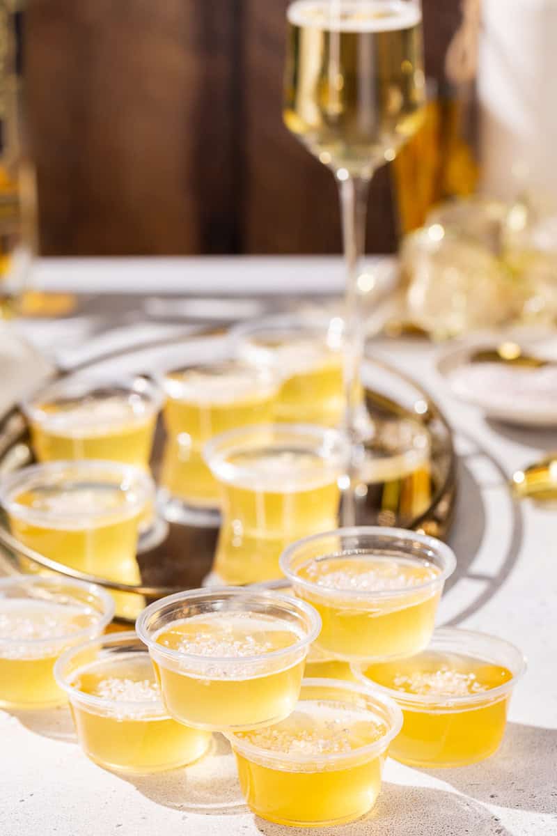 Mirrored tray carrying Champagne jello shots with gold New Year’s celebration decor in the background. Some of the jello shots are on the countertop.