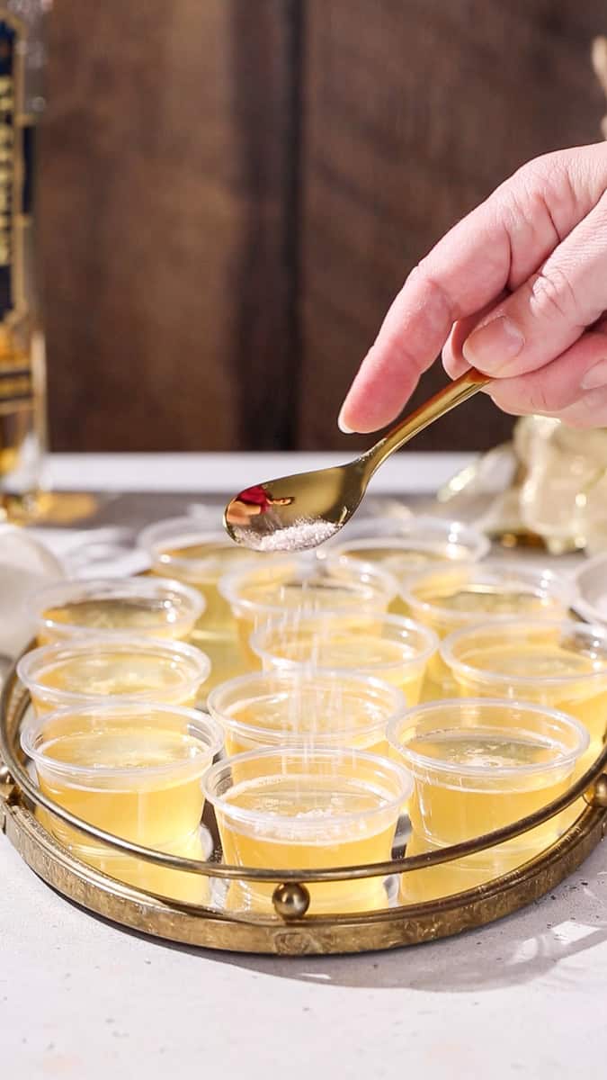 Hand using a spoon to add silver sugar to the top of some Champagne jello shots after they chilled and set.