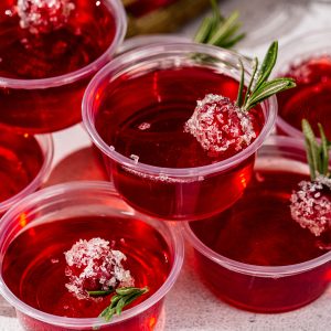 Close up of a stack of cranberry Christmas jello shots, each with a sugared cranberry and rosemary garnish.