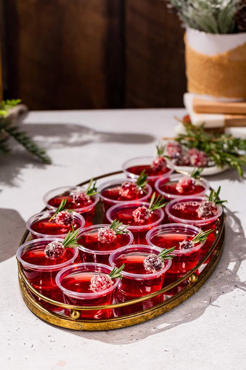 A tray filled with Cranberry Christmas Jello Shots on a countertop. There are candied cranberries in the background along with some holiday decor.