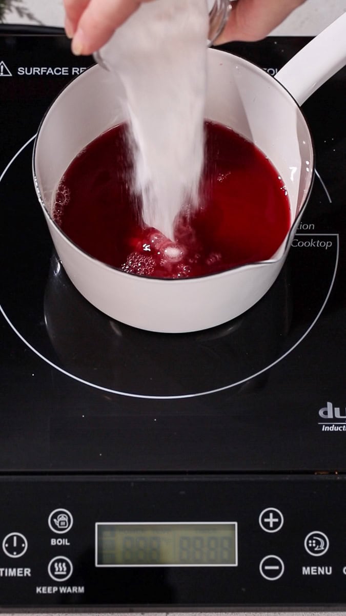 Hand pouring sugar into a saucepan filled with red liquid.