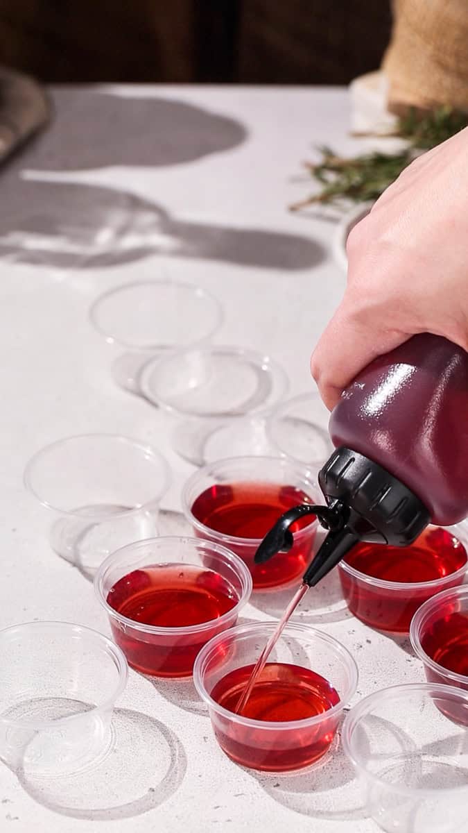 Hand using a squeeze bottle to add red liquid to mini shot cups.