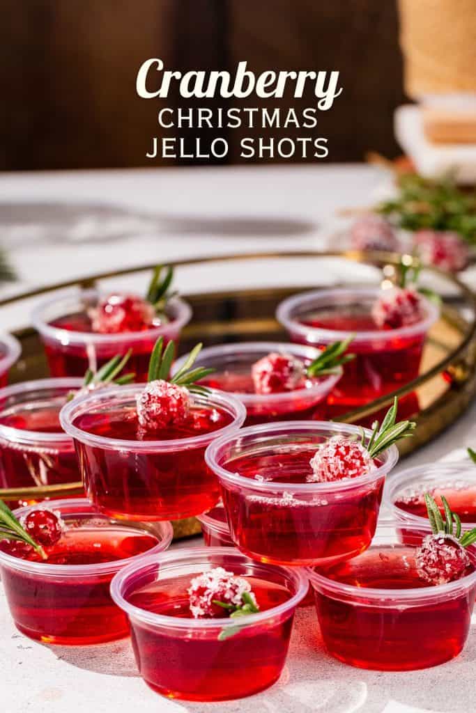 Cranberry Christmas Jello Shots in a group together on a countertop. Fresh rosemary and cranberry are in the background along with holiday decor. Text overlay above the shots says “Cranberry Christmas Jello Shots” in all caps.