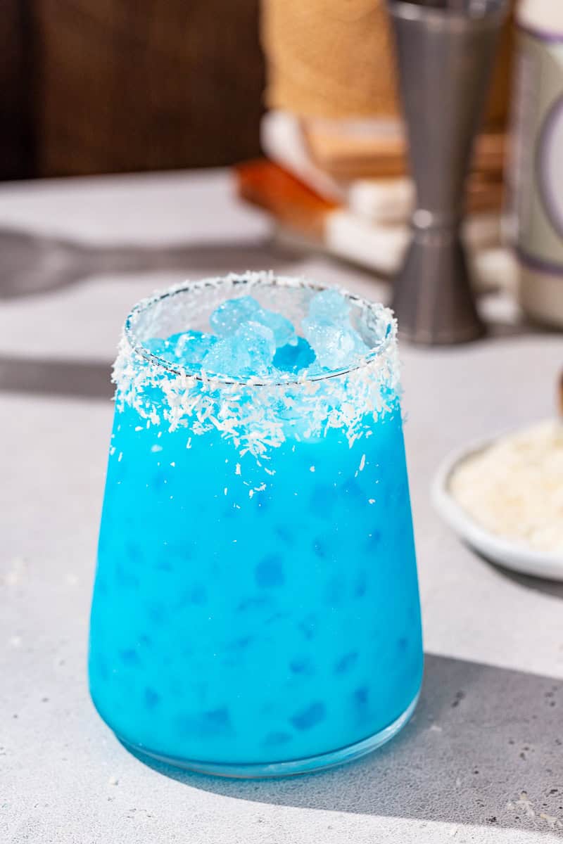 Jack Frost cocktail on a countertop with some bar tools and a dish of dried coconut in the background.