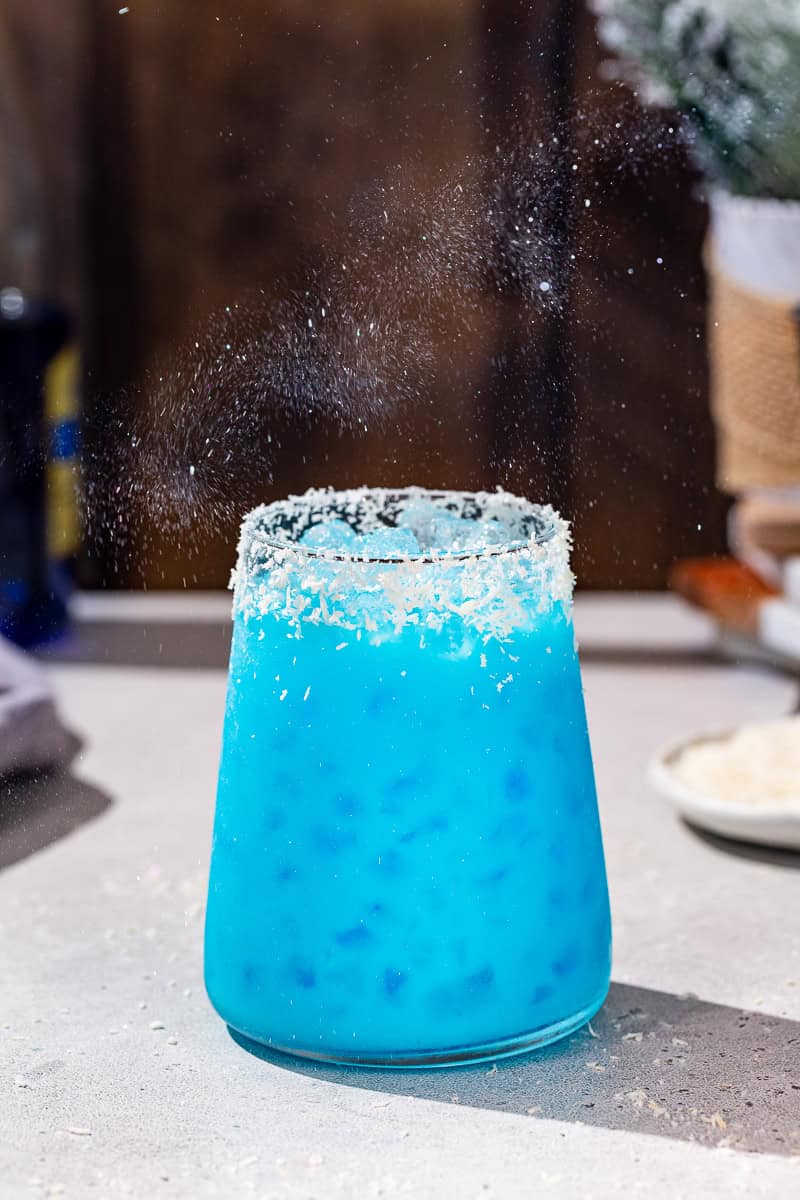 Side view of a blue colored Jack Frost cocktail on a countertop with edible glitter floating in the air above it.