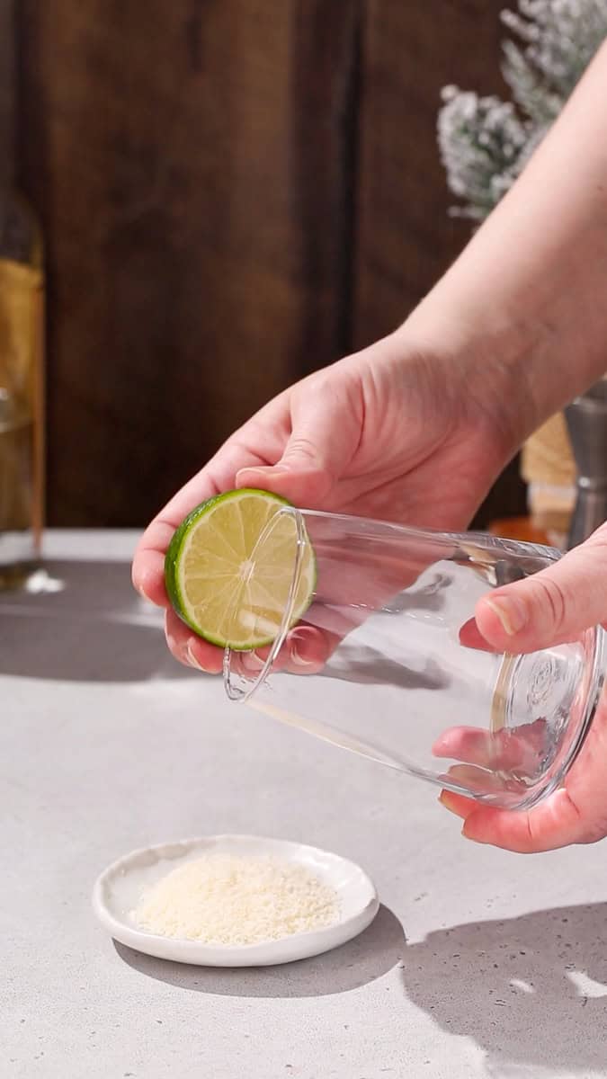 Hands using a cut lime to wet the rim of a cocktail glass.