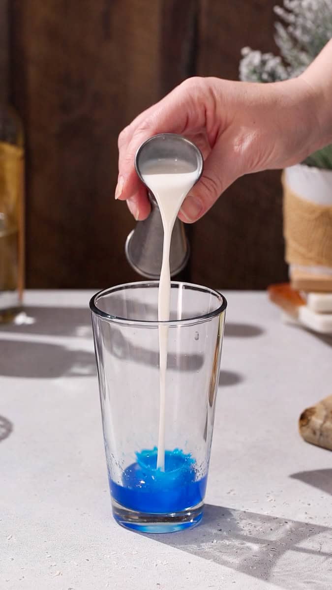 Hand adding cream of coconut to a cocktail shaker filled with blue liquid.