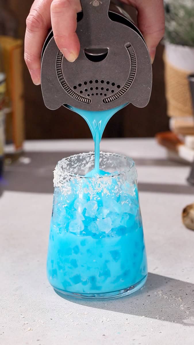 Hand straining a bright blue cocktail into a cocktail glass filled with nugget ice.