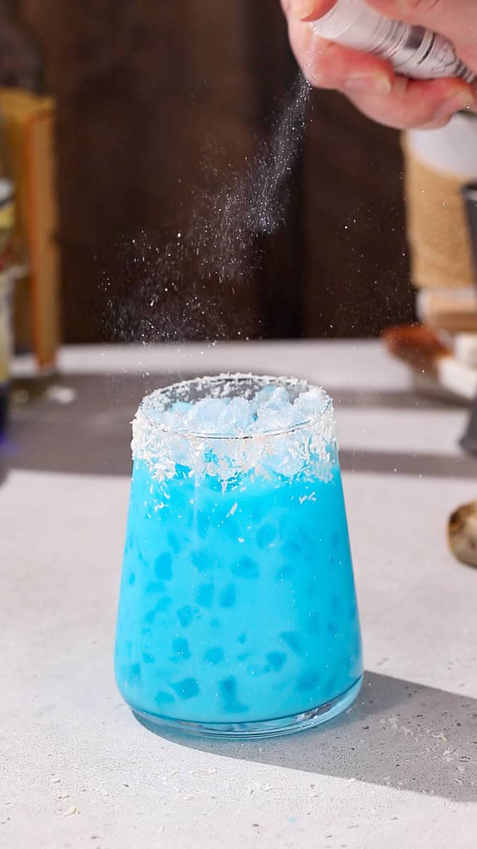 Hand using a pump bottle to add edible glitter to the top of a bright blue drink.