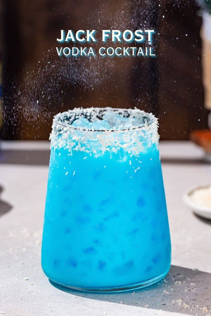 Side view of a blue colored Jack Frost cocktail on a countertop with sparkle glitter in the air above it. Text overlay says “Jack Frost vodka cocktail”.