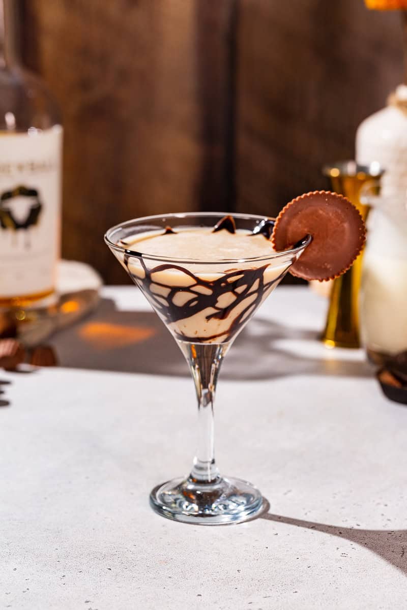 Side view of a Peanut Butter Cup Martini cocktail in a stemmed martini glass. The glass has a chocolate drizzle and there is a peanut butter cup candy garnish.