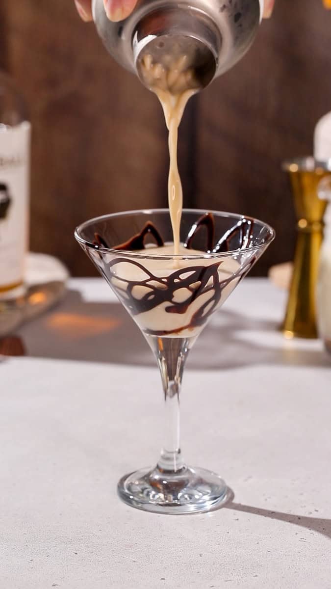 Hand straining a drink into a chocolate-drizzled martini glass.